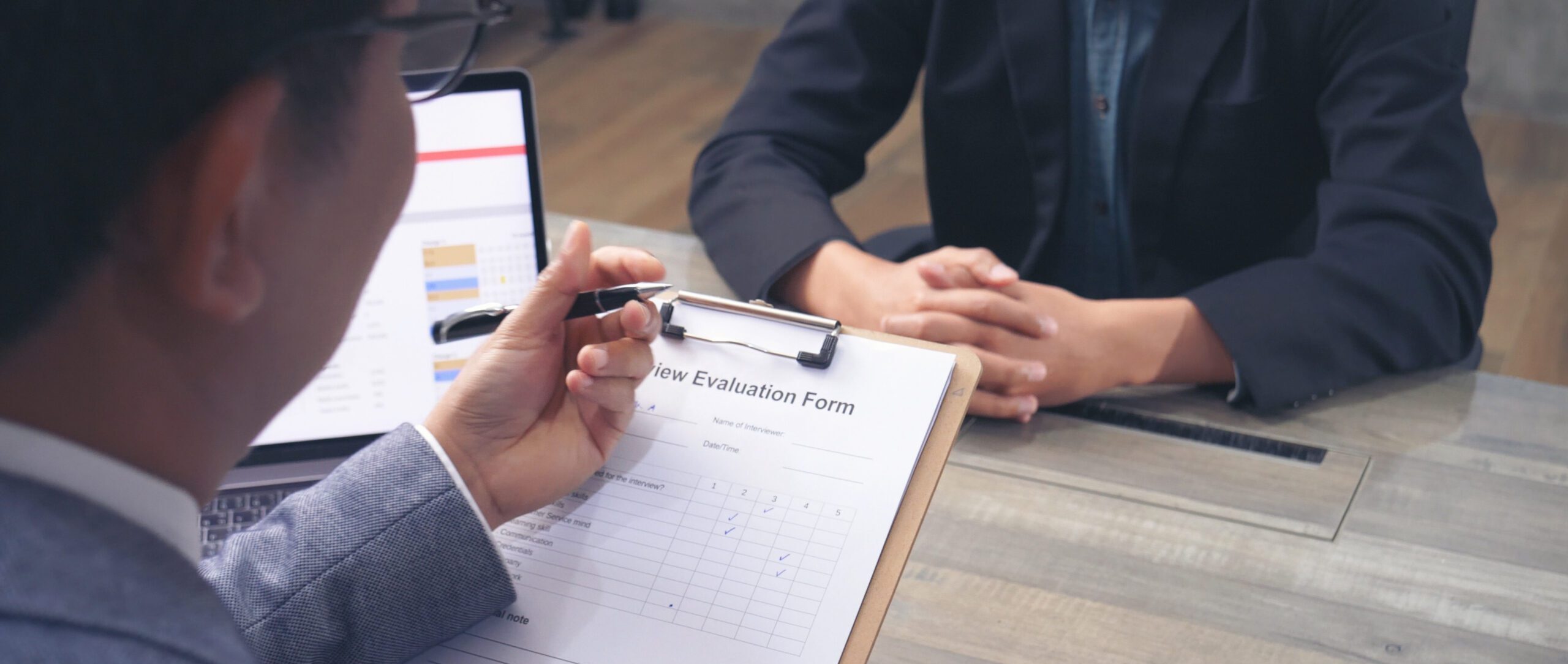 Person conducting a job interview, holding a clipboard with an interview evaluation form, while the candidate sits across the table with hands folded in a professional office setting.